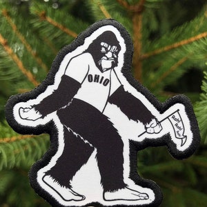 May include: Embroidered patch depicting a black and white Bigfoot. The Bigfoot is wearing a white jersey with "OHIO" on it and holding a flag that says "Rah!". The patch has a white border and is set against a green background.