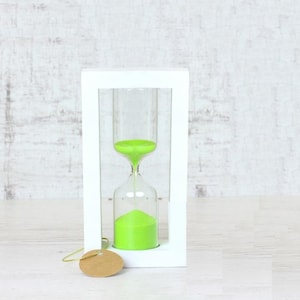 Puede incluir: Un reloj de arena blanco con arena verde dentro de un marco de madera blanco. El reloj de arena está sobre una superficie blanca.