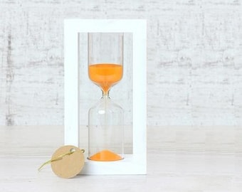 Könnte beinhalten: Eine weiße Sanduhr mit orangefarbenem Sand im Inneren, die auf einem weißen Holzsockel steht. Die Sanduhr befindet sich in einem weißen Rahmen.