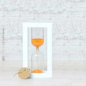 May include: A white hourglass with orange sand inside, sitting on a white wooden base. The hourglass is in a white frame.