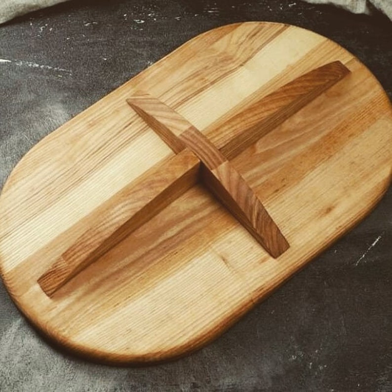 May include: A wooden serving board with a natural finish. The board is oval shaped and has a cross-shaped design in the center.