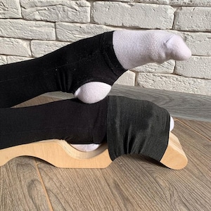 以下が含まれることがあります： 木製の足ストレッチ器具に、黒い布のストラップと白い靴下を履いた足が乗っています。足はストレッチ器具の上に置かれており、器具は木製の床の上にあります。