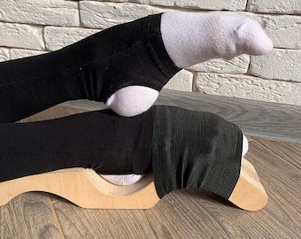 May include: A wooden foot stretcher with a black fabric strap and a white sock on the foot. The foot is resting on the stretcher, which is on a wooden floor.