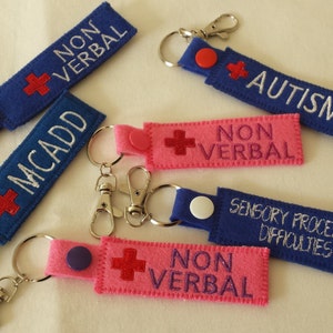 Op de afbeelding: Zes verschillende sleutelhangers met een rood kruis en tekst. De sleutelhangers zijn gemaakt van blauw en roze vilt. De tekst op de sleutelhangers is "NON VERBAL", "MCADD", "AUTISME", "SENSORISCHE VERWERKINGSPROBLEMEN" en "NON VERBAL".