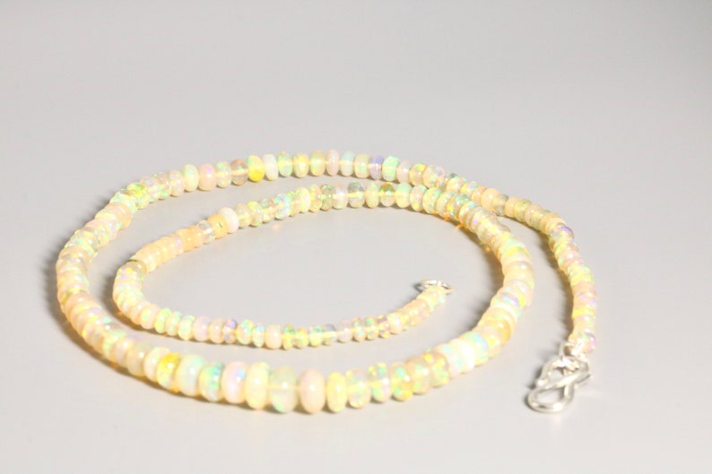 May include: A long necklace made of small, round, iridescent opal beads. The necklace is light yellow in color and has a silver clasp.