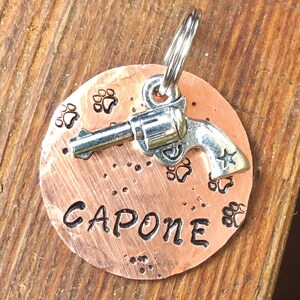 May include: Copper dog tag featuring a silver gun charm and the name "CAPONE" engraved on it. The tag is decorated with paw prints.