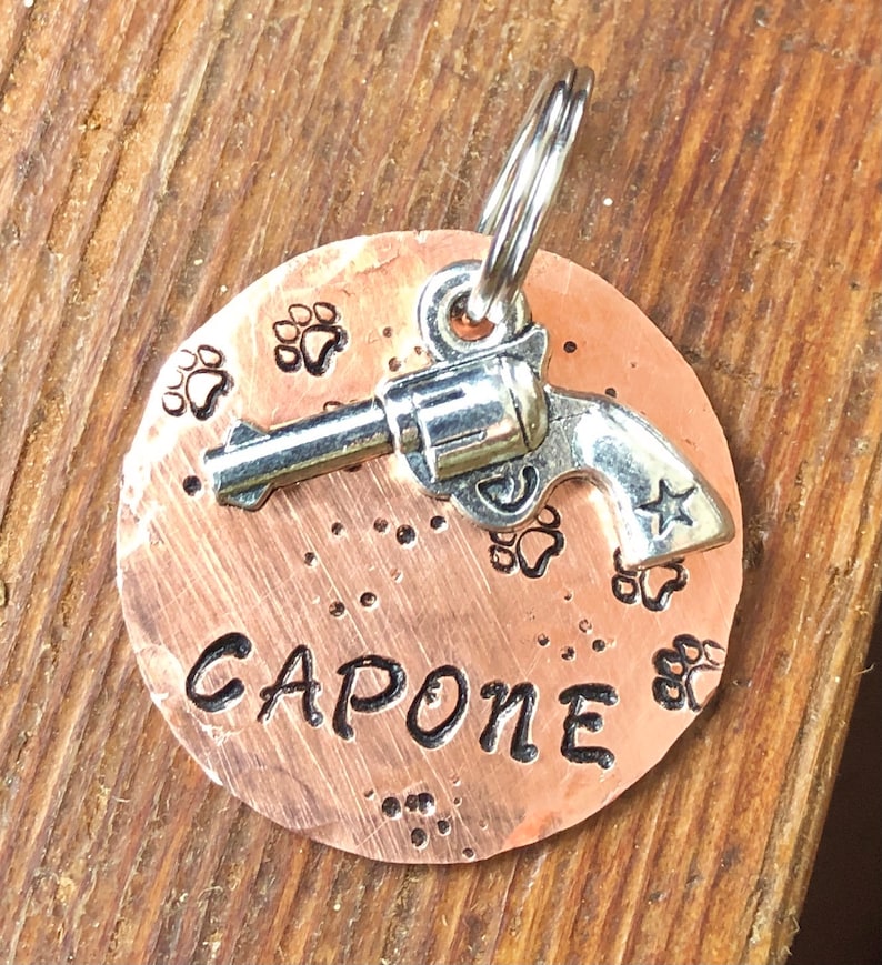 May include: Copper dog tag featuring a silver gun charm and the name "CAPONE" engraved on it. The tag is decorated with paw prints.