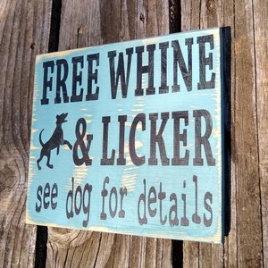 May include: A rustic wooden sign with a distressed blue paint finish. The sign reads "FREE WHINE & LICKER see dog for details" with a black silhouette of a dog in the upper left corner.