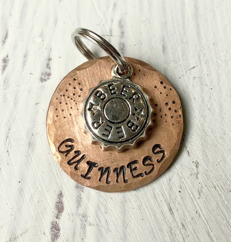 Pu&ograve; includere: Un'etichetta per cani in rame con un ciondolo a forma di tappo di bottiglia in argento che dice "BEER" e il testo "GUINNESS" inciso sull'etichetta.