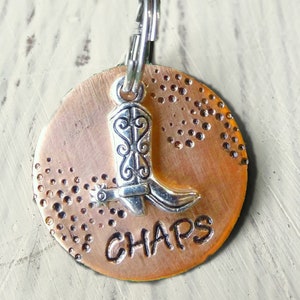 May include: A copper-colored metal tag with a silver cowboy boot charm and the word "CHAPS" engraved on it. The tag has a decorative pattern of small dots.