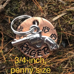 May include: A copper dog tag with a silver gun charm and paw prints. The tag is engraved with the name "RUGER" and measures 3/4 inch in diameter.