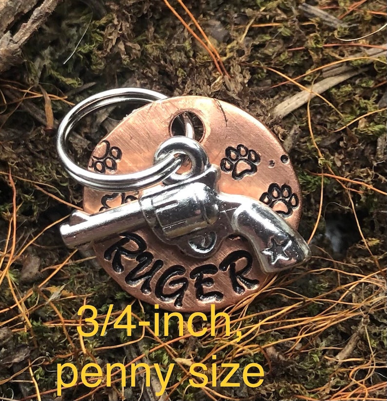 May include: A copper dog tag with a silver gun charm and paw prints. The tag is engraved with the name "RUGER" and measures 3/4 inch in diameter.