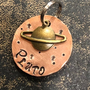 May include: A copper dog tag with a gold-toned planet charm and the word "PLUTO" engraved on it. The tag has a silver ring for attaching to a collar.