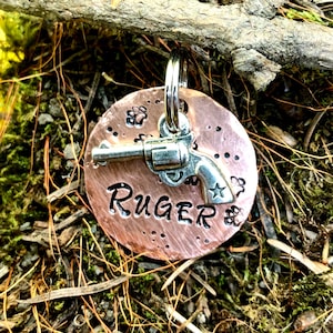 May include: Copper dog tag featuring a silver revolver and the name "RUGER" engraved on it.