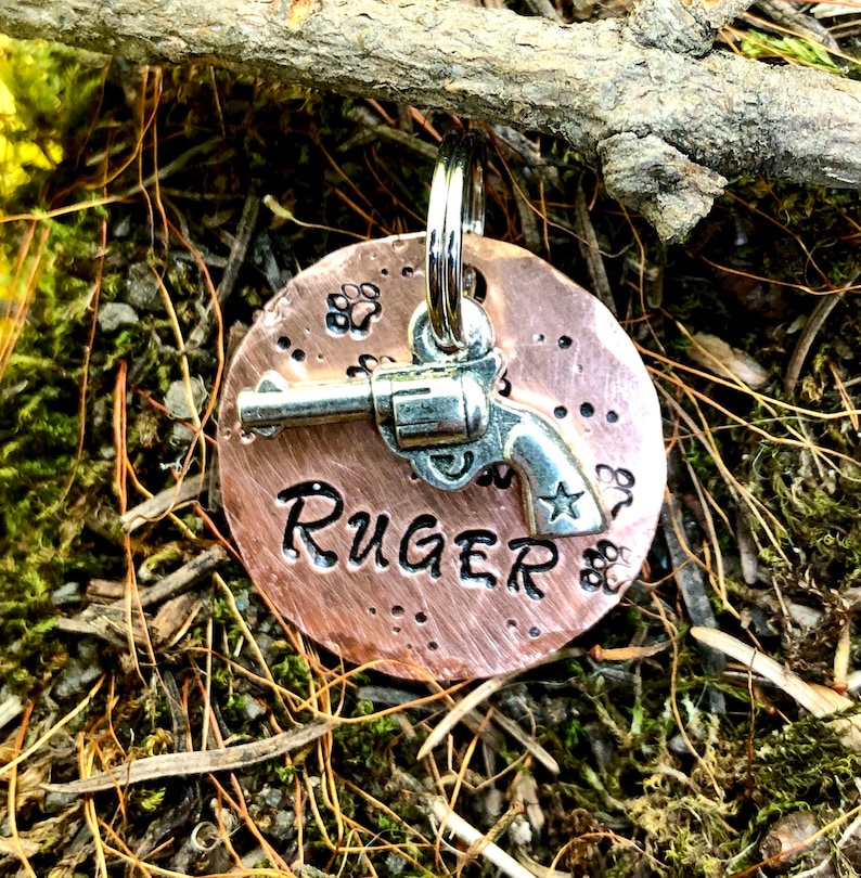 May include: Copper dog tag featuring a silver revolver and the name "RUGER" engraved on it.