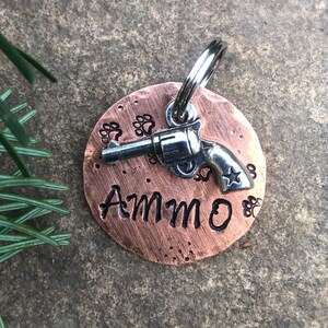 May include: Copper dog tag featuring a silver gun charm and the name "AMMO" engraved on it. The tag is surrounded by paw prints.