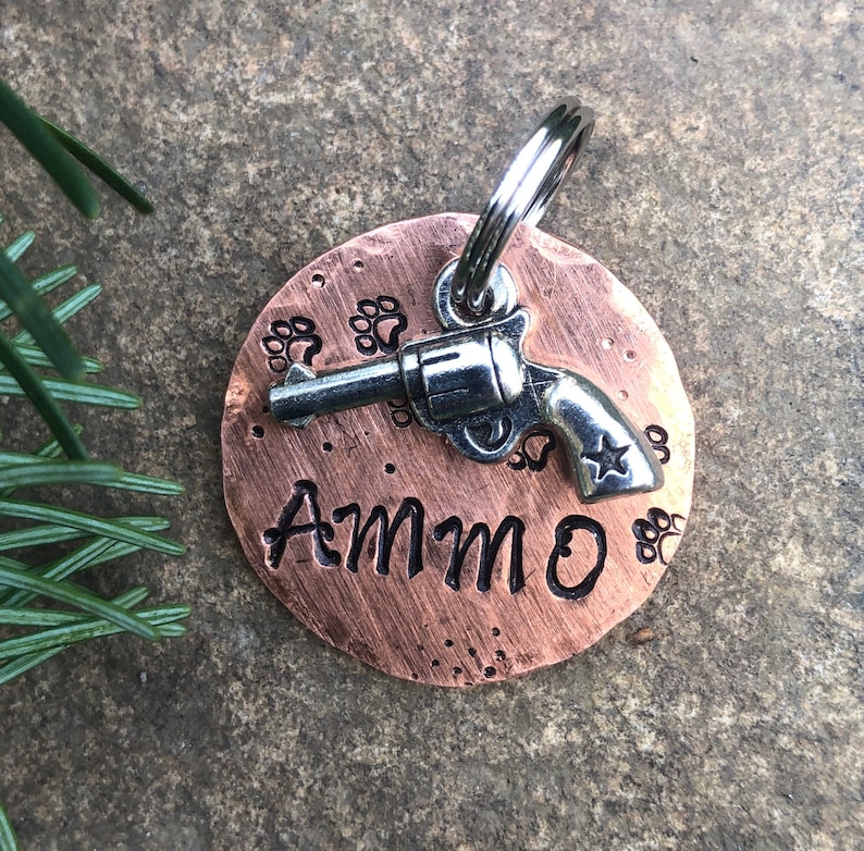 May include: Copper dog tag featuring a silver gun charm and the name "AMMO" engraved on it. The tag is surrounded by paw prints.