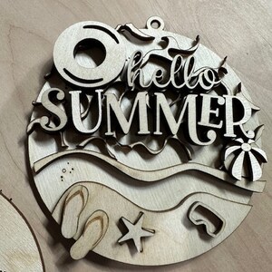 May include: Wooden cutout of a beach scene with the words "hello summer" in the center. The scene includes a life preserver, flip flops, a starfish, a diving mask, and a beach ball.