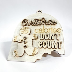 May include: A light-coloured wooden ornament shaped like a dome with a bite taken out of the bottom. It features a layered gingerbread man figure and the engraved text "Christmas calories DON'T COUNT" with a small hole at the top for hanging.