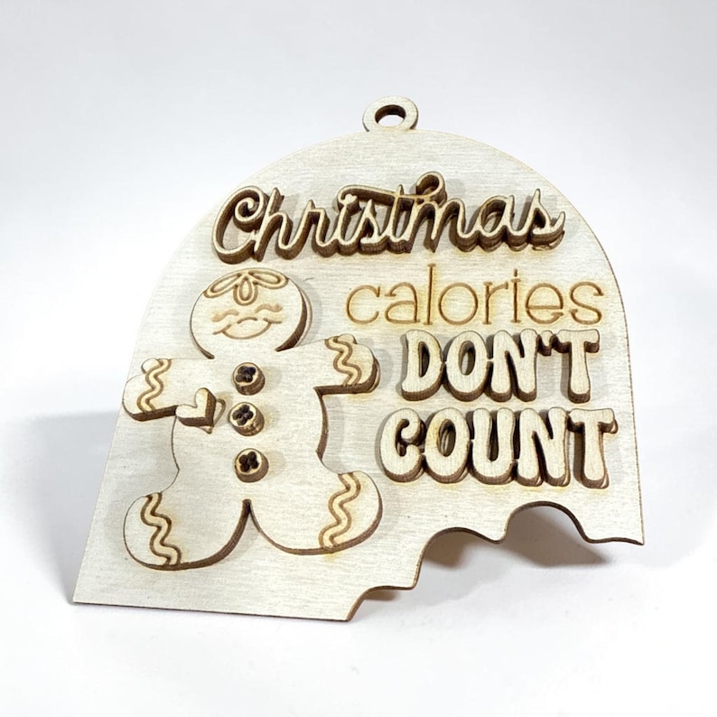 May include: A light-coloured wooden ornament shaped like a dome with a bite taken out of the bottom. It features a layered gingerbread man figure and the engraved text "Christmas calories DON'T COUNT" with a small hole at the top for hanging.