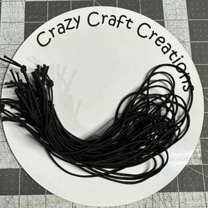 May include: A close-up of a pile of black elastic string on a white circular surface with the text "Crazy Craft Creations" printed on it.