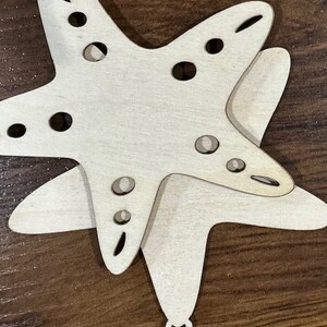 May include: A wooden cutout of a starfish with a hole in the center for hanging. The starfish is painted white and has a natural wood grain.
