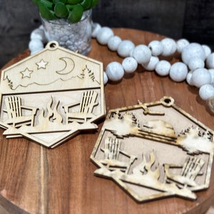 May include: Two wooden ornaments shaped like hexagons with a campfire scene. One ornament features a crescent moon and stars, while the other has a sun. Both ornaments depict two chairs and a campfire.