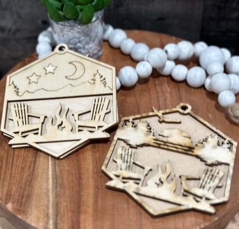 May include: Two wooden ornaments shaped like hexagons with a campfire scene. One ornament features a crescent moon and stars, while the other has a sun. Both ornaments depict two chairs and a campfire.