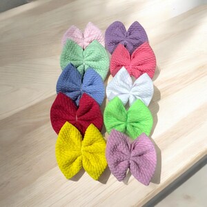 May include: A set of 12 colorful hair bows made of textured fabric. The bows are arranged in a row, with the colors ranging from light pink to purple.