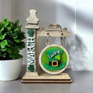 May include: Wooden sign with a green shamrock hat and the word "Lucky" on a green circle. The sign is hanging from a wooden post with the word "MARCH" on it.