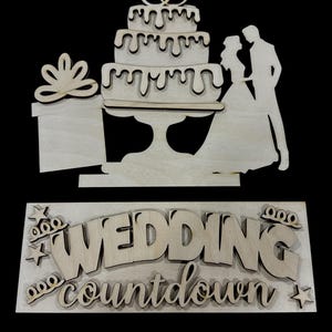 May include: Wooden wedding cake topper with a three-tiered cake, rings, and a bride and groom silhouette. Includes a gift box and a sign that reads "WEDDING countdown" with stars and crowns. Perfect for wedding decor.