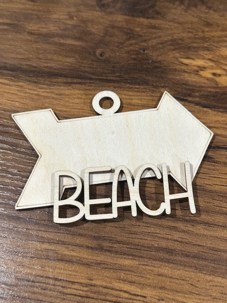 May include: Wooden arrow sign with the word "Beach" in white lettering. The sign is unfinished and ready to be painted or stained.