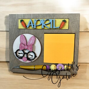 May include: A wooden sign with the word "APRIL" in blue letters on a yellow background. The sign also features a bunny face with pink ears and glasses, a yellow sticky note, and a brown pen with purple and yellow beads.