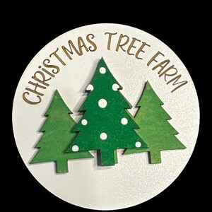 May include: A white round wooden sign with the text "Christmas Tree Farm" in gold lettering. Three green Christmas trees with white polka dots are layered on top of the text.