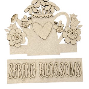 May include: A wooden spring-themed decoration featuring a watering can shape with flowers, bees, and a heart. The words "SPRING BLOSSOMS" are displayed on a separate wooden piece below the watering can.