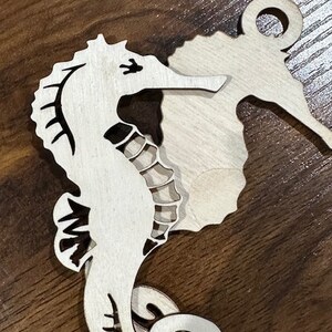 May include: A wooden cutout of a seahorse with a decorative swirl design. The seahorse is painted white and has black details.