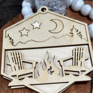 May include: Wooden cutout of a campfire scene with two chairs and a fire. The cutout is in the shape of a hexagon and has a moon, stars, and trees in the background.