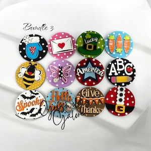 May include: A set of 12 round wooden signs with various designs and sayings. The signs feature polka dots and include designs such as a bee, butterfly, a heart in an envelope, a snowman, a leprechaun, carrots, ABCs, a pencil, bats, a turkey, a Santa Claus, and the words "Give Thanks" and "Hello Fall".