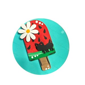 May include: A round wooden sign with a turquoise background features a watermelon popsicle with a white flower and a black ant. The popsicle is painted red with black seeds and a green stripe. The stick is brown with white dots.