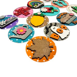 May include: A set of 12 wooden round ornaments with various seasonal themes. The ornaments feature a variety of colors, including red, blue, green, yellow, orange, and white. Each ornament has a different design, such as a snowman, a ghost, a pumpkin, a lemon, a watering can, a rainbow, and a maple leaf.