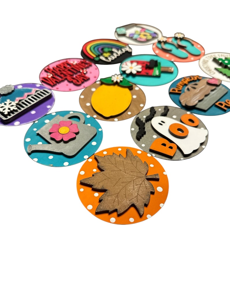 May include: A set of 12 wooden round ornaments with various seasonal themes. The ornaments feature a variety of colors, including red, blue, green, yellow, orange, and white. Each ornament has a different design, such as a snowman, a ghost, a pumpkin, a lemon, a watering can, a rainbow, and a maple leaf.