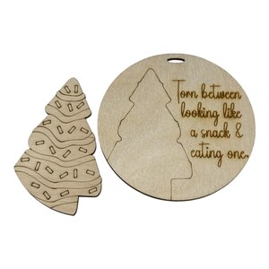 May include: Two wooden Christmas tree ornaments. One is shaped like a tree with icing and sprinkles. The other is a circle with a tree cutout and the text "Torn between looking like a snack & eating one."