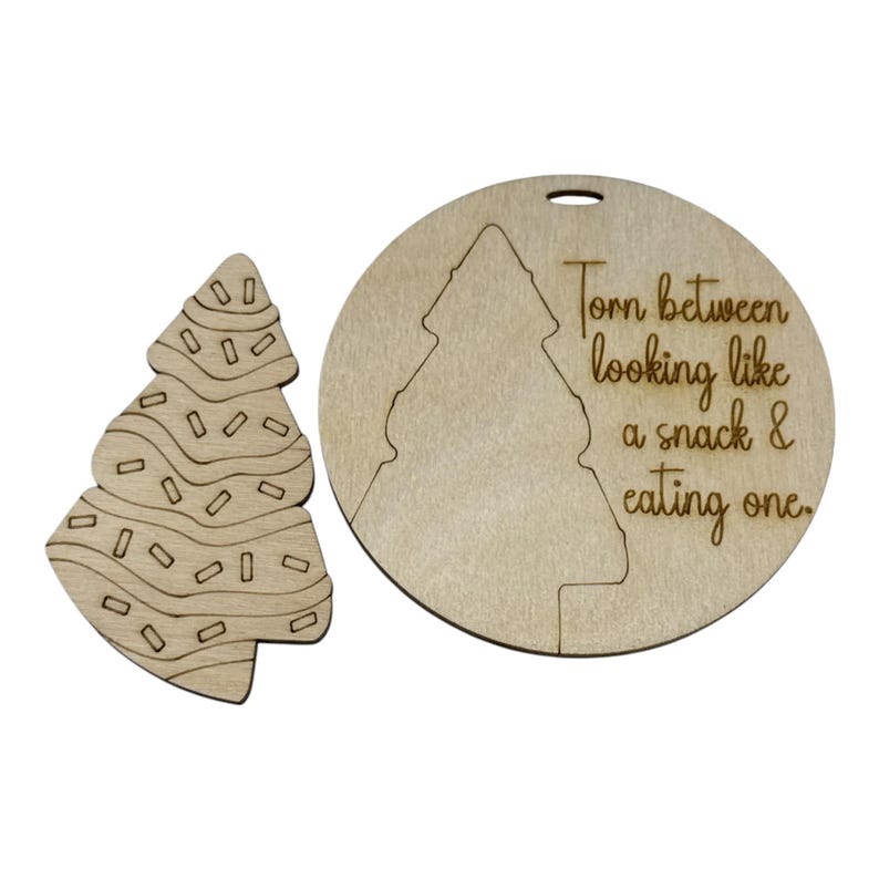 May include: Two wooden Christmas tree ornaments. One is shaped like a tree with icing and sprinkles. The other is a circle with a tree cutout and the text "Torn between looking like a snack & eating one."