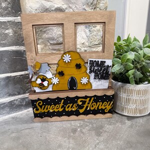 May include: Wooden decorative piece with a bee theme. Features a honey jar, a beehive, and a sign that says "Home Sweet Home". The base reads "Sweet as Honey" in yellow script on a black background. A small potted plant is to the right.