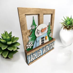 May include: Wooden window cutout with a snowman and pine trees inside. The window has a blue and white polka dot border and the text "Hello Winter" on the bottom.