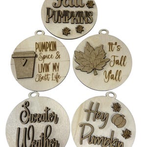 May include: Five wooden fall-themed ornaments. The ornaments are round with various fall-related phrases and images, including "Fall Pumpkins," a coffee cup, a maple leaf, and a pumpkin. Each ornament has a small hole for hanging.