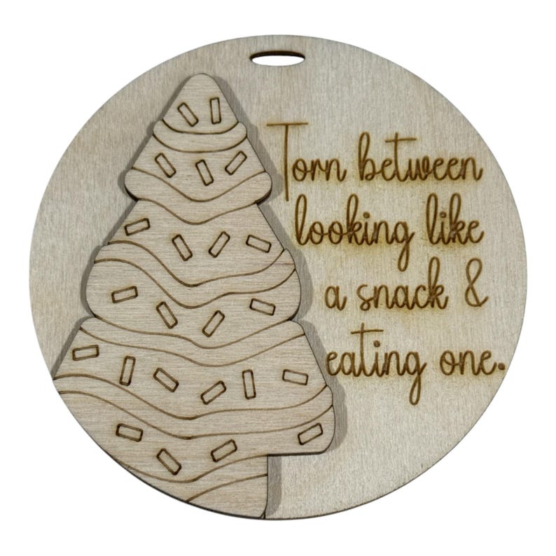 May include: A round, wooden Christmas ornament with a laser-cut design of a Christmas tree and the phrase "Torn between looking like a snack & eating one." The ornament has a hole for hanging.