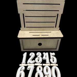 May include: A wooden card holder with a slot and a drawer, accompanied by white numbers 0-9 with stitched detailing. The holder has a curved top and horizontal slats. The numbers are designed to stand upright.