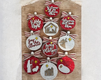 Puede incluir: Doce adornos navideños redondos, rojos y blancos con diversas ilustraciones y textos religiosos, incluyendo "O Holy Night", "Wisemen Still Seek Him", "Away in a Manger" y "Joy to the World".