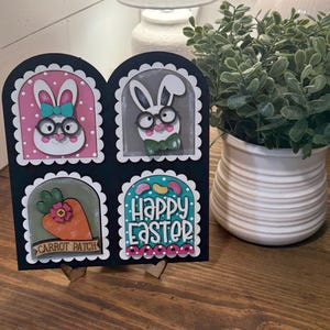 May include: A black Easter-themed wooden decoration featuring four panels. Two panels depict bunny faces wearing glasses, one shows a carrot patch, and the last reads "Happy Easter." The decoration is supported by two wooden stands.